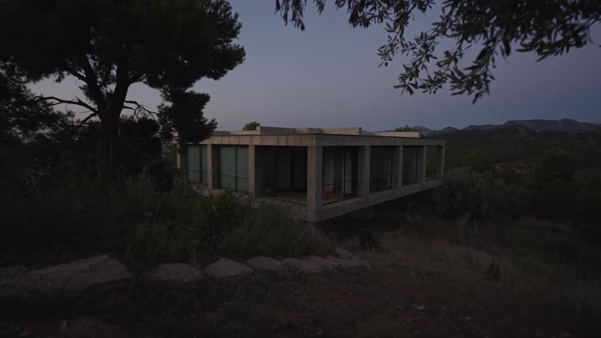 Modern concrete house with large glass windows set among vegetation and hills at dusk