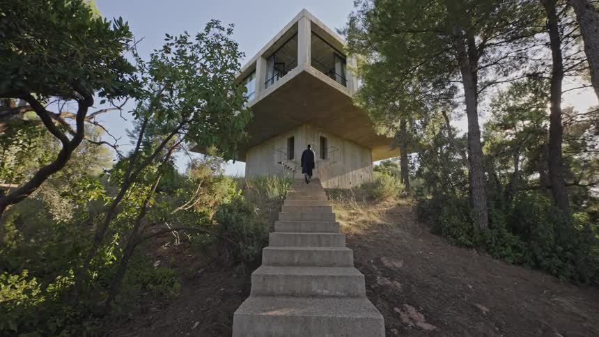 A man in dark clothing walks up a set of concrete stairs toward a modern geometric building with large windows on a grassy hill surrounded by green trees