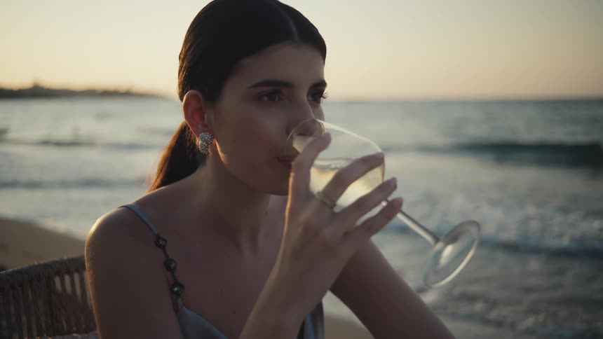 A woman in a satin dress sips wine by the shore as the evening sun sets over the ocean