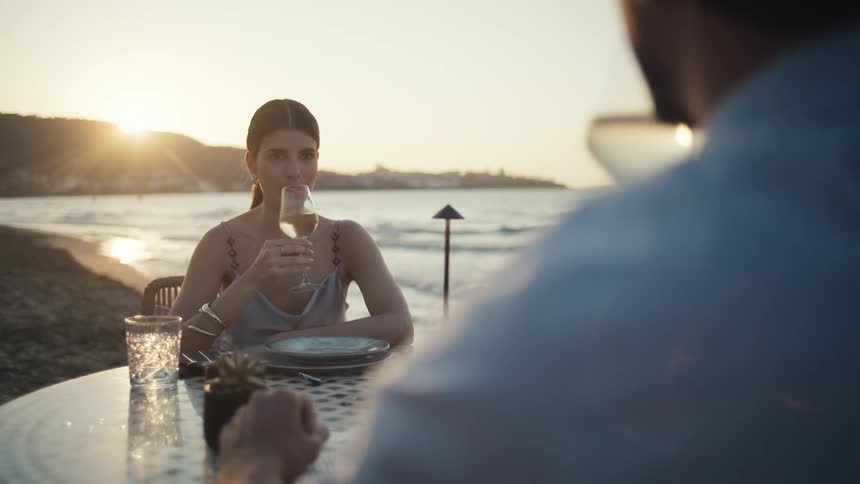 A woman and a man sit at a small table on a beach and the woman raises her wine glass to toast while the sun sets over the ocean behind her