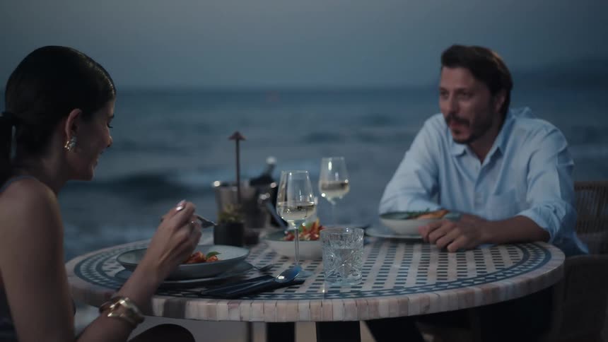 A couple is enjoying a romantic dinner by the sea at night with the woman playfully feeding the man a spoonful of food