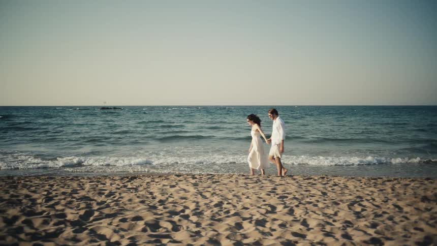 The video shows a couple holding hands and walking on a sandy beach they are both dressed in white with the woman wearing a dress and the man wearing a shirt and shorts