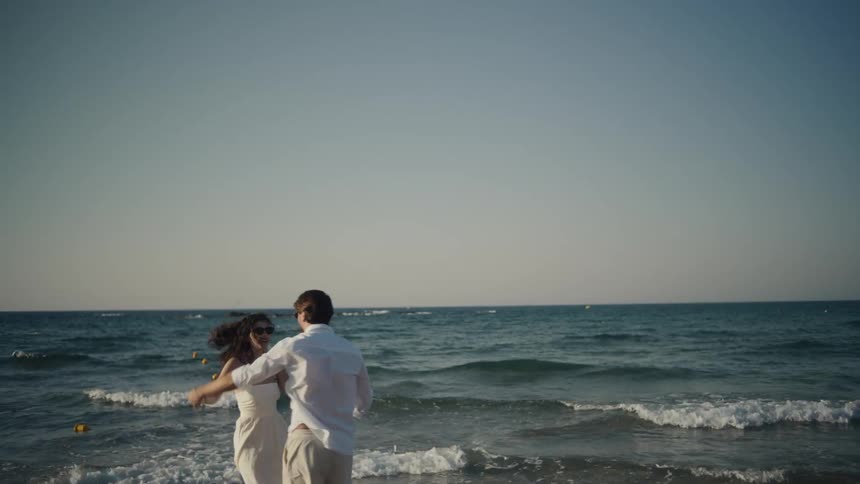 A couple holds hands and dances while standing barefoot in the gentle surf at sunset