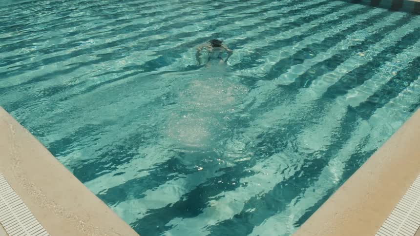 A highangle shot captures a person wearing a black swimsuit as they dive headfirst into a swimming pool creating a splash and then swimming away underwater