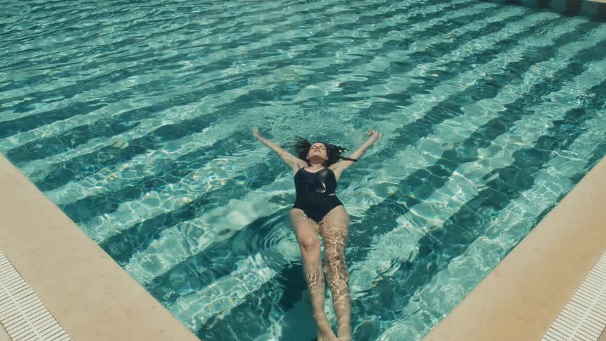 A highangle shot shows a woman wearing a black swimsuit relaxing on her back and floating serenely in a swimming pool on a sunny day