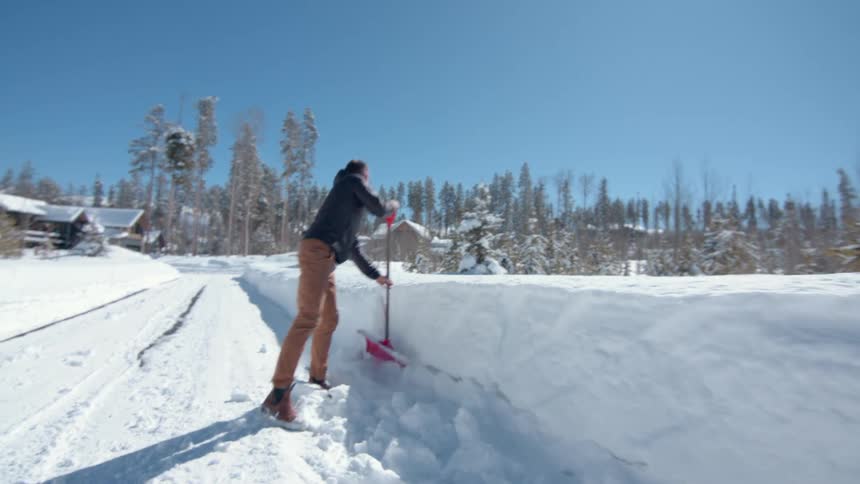 The video shows a man standing in a snowcovered landscape using a red shovel to clear a path he is wearing sunglasses and is surrounded by high banks of snow and a forest of trees