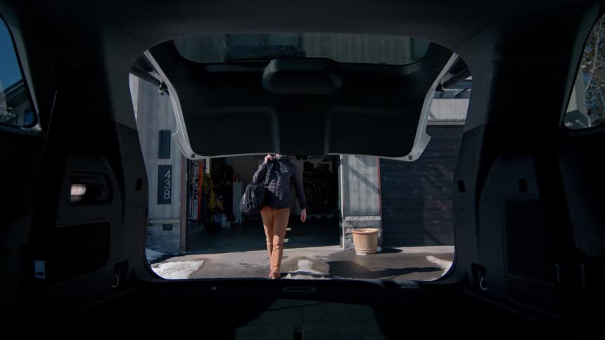 A shot from inside the rear of a vehicle shows a man walking towards the car with a duffel bag preparing to load it into the trunk