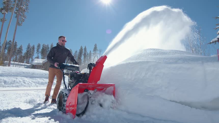 A man wearing sunglasses and a black jacket pushing a snow plow along the side of the road at midday clearing snow to the roadside