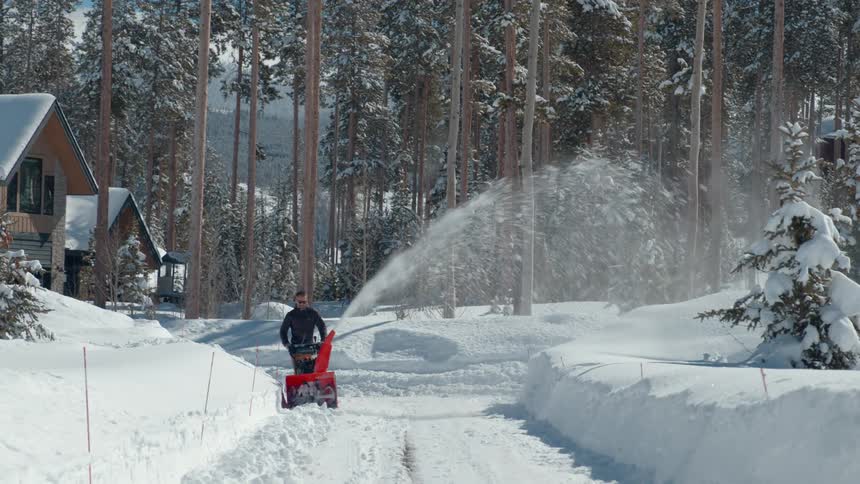 A man uses a snow blower to clear a driveway in a snowy forest neighborhood