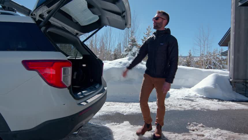 A young man walks out of a garage carrying a bag puts it in the trunk of a car gets in and drives off on a snowy road at midday