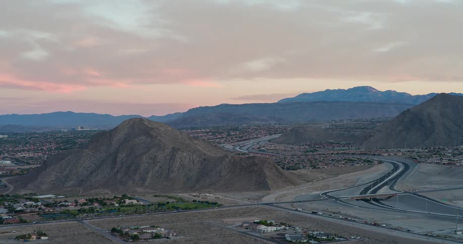 An aerial view of a winding highway and surrounding buildings nestled in a desert valley with large barren mountains in the background under a pastelcolored sunset sky