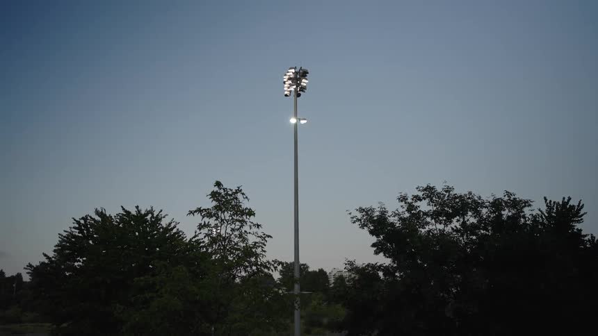 A sports field with a tall spotlight turned on at evening with a row of trees behind it