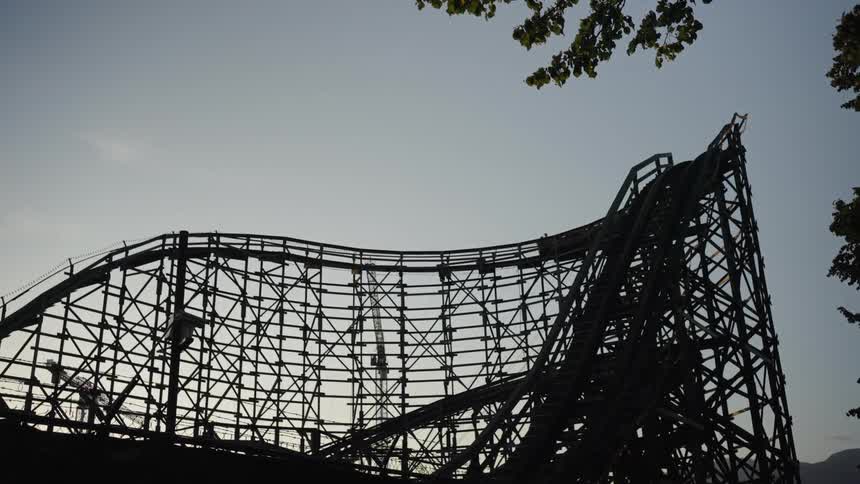 A silhouette of a large wooden rollercoaster is shown against a pale sky the rides car is seen moving up a steep incline and then descending down the other side