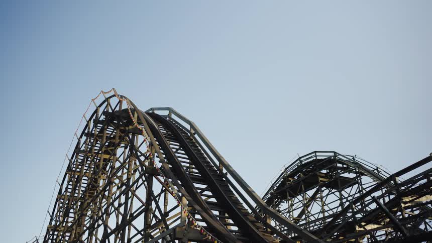 A towering wooden roller coaster rises and dips sharply against a clear blue sky