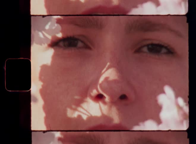 A woman with a serious facial expression looks directly at the camera through a frame of blurry pink and white flowers with the sunlight casting a strong shadow on her face as she looks down and back up again