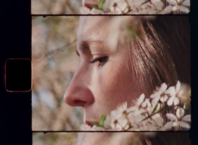 Sideways portrait or profile of a brown-haired woman standing next to flowers on a branch covering her face as she enjoys the warm sunlight while looking down in a contemplative and melancholic mood