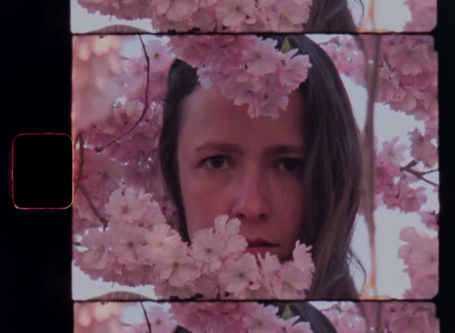 Young woman standing under tree in full bloom with pink flowers turning her head with long brown hair looking straight at the vintage film camera staring at the viewer with determined and self-confident expression
