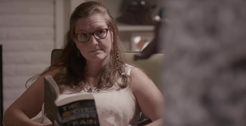 A woman sits in a chair and reads a book before being helped up by an outstretched hand