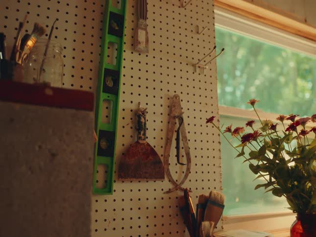 Sunlight streams through a window onto a pegboard wall of tools fresh flowers in a brown vase and scattered paintbrushes