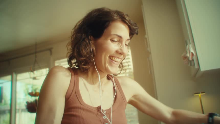 A young woman in the kitchen doing her chores while wearing wired earbuds singing and having fun