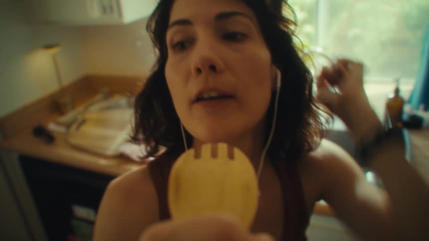 A female is singing in her kitchen into a wooden spoon while wearing ear buds