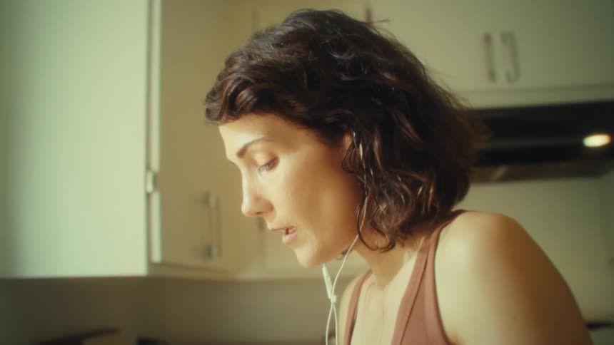 Wearing earphones a woman gestures expressively while singing in a sunlit kitchen with white cabinets and a teal kettle