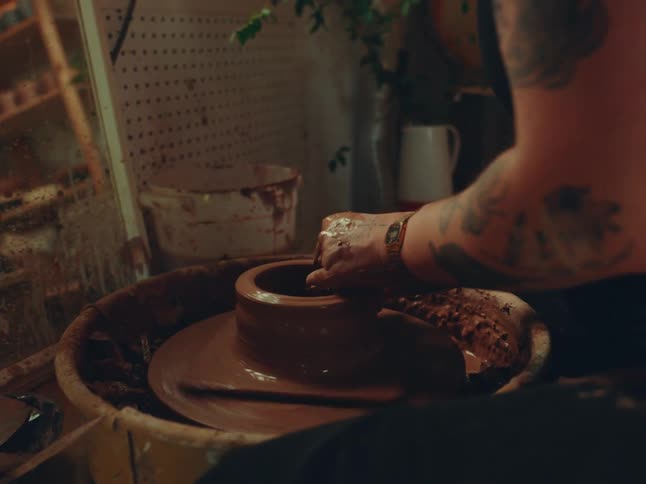 A young woman using a pottery wheel to make a clay vase with a mirror in front of her inside a studio
