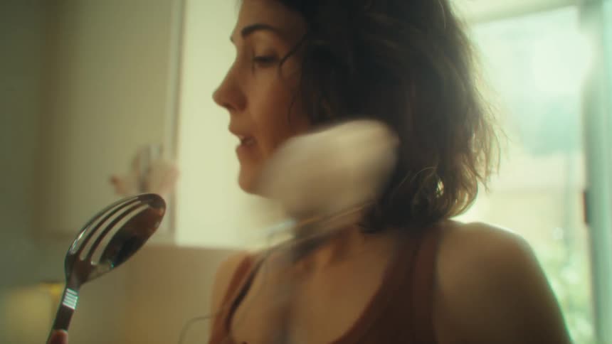 A young woman uses a stainless steel spoon as a microphone and sings into it wearing wired earbuds and having a fun time in the kitchen at midday