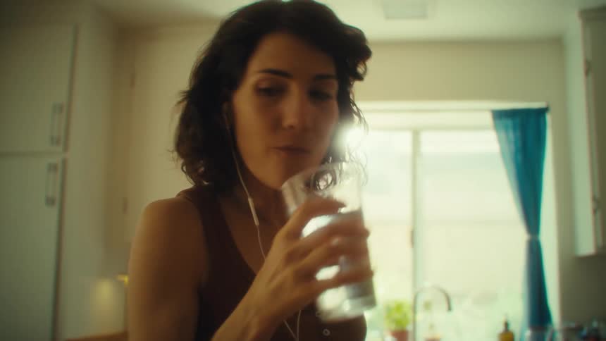 The video shows a woman with dark hair and headphones in her ears standing in a kitchen she takes a sip of water from a clear glass and seems to be enjoying the music she is listening to