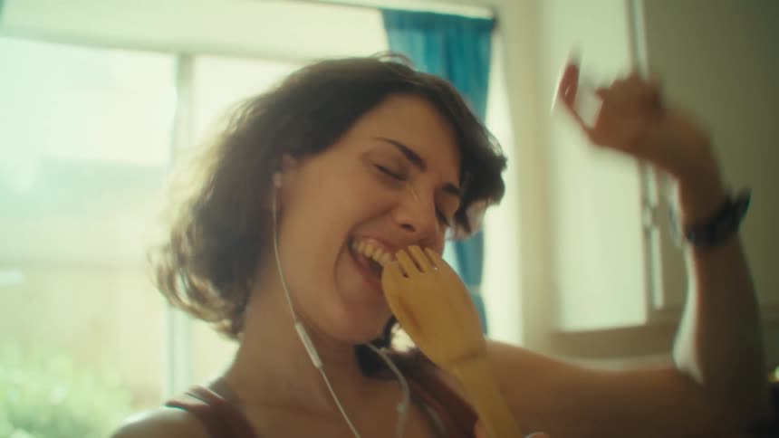 A woman with long brown hair smiles and sings into a wooden spoon as she wears earbuds and dances in a kitchen with a window and a blue curtain behind her