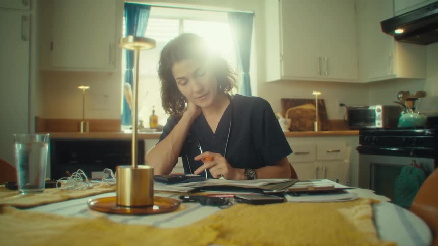 A woman in scrubs studies paperwork and uses a calculator at a sunlit kitchen table with a stethoscope draped around her neck