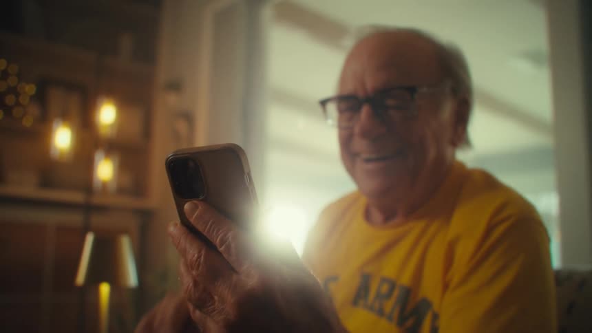 Elderly man wearing glasses smiles while holding a phone with both hands