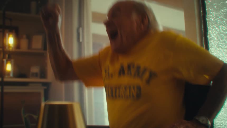 Elderly man in a wheelchair smiles broadly wearing a yellow army veteran shirt