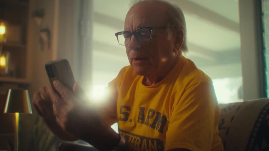 Elderly man wearing glasses looks at the phone he holds with both hands