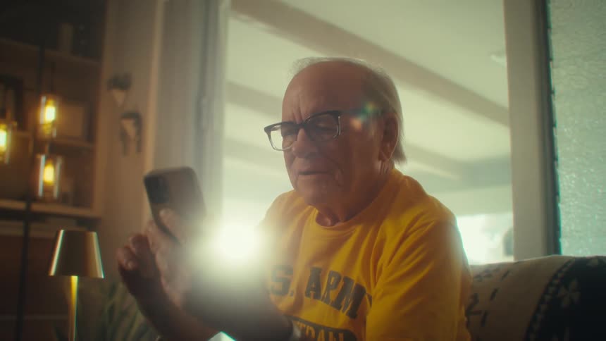 An elderly man scrolls and laughs as he uses a mobile phone while sitting in a living room during daytime