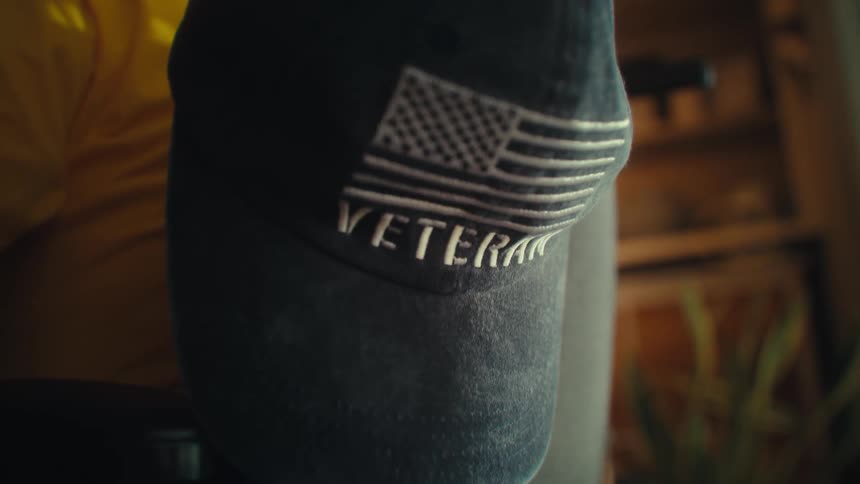 A closeup shot of a persons shoulder with an american flag patch and a veteran patch on a sleeve is shown the person is wearing a dark blue shirt and is standing in a dimly lit room with a light source to their side