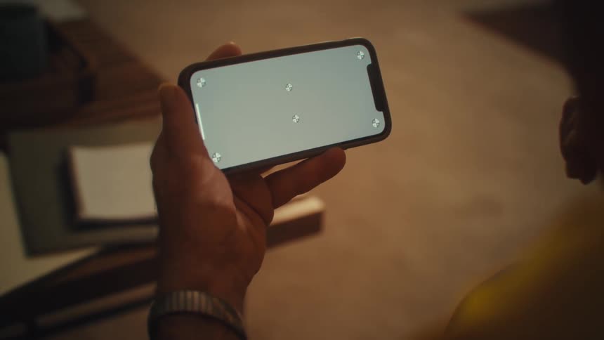 A man holding a phone and wearing a wristwatch sitting in the living room watching content on his device in front of him is a coffee table cluttered with books and documents
