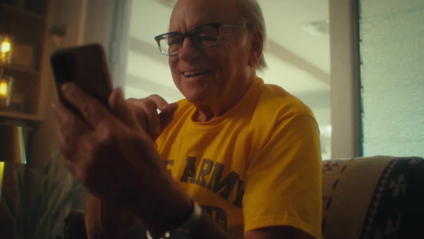 An older man wearing a yellow us army veteran shirt smiles while looking at his phone in a sunlit room