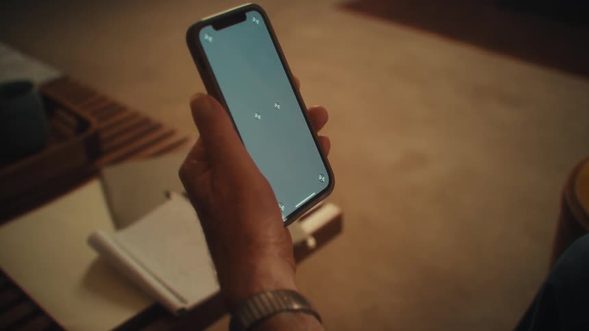 A man holding his smartphone sits indoors with a coffee table in front of him covered in documents
