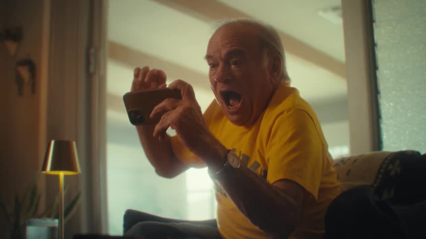Elderly man in a yellow tshirt smiles excitedly while watching something on his cell phone