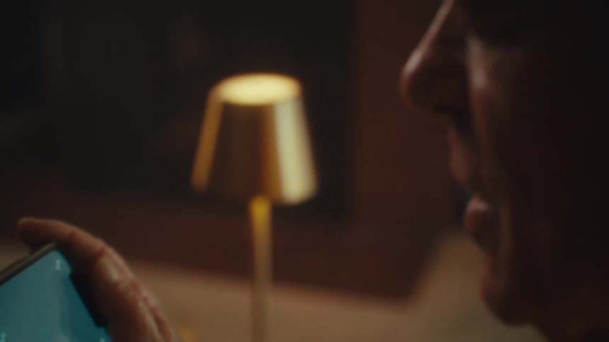 An elderly man smiles and laughs while looking at his phone in a dimly lit room