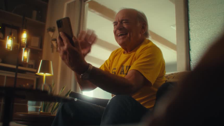 An elderly man sits in his living room smiling and holding his phone while waving to the caller during a video call