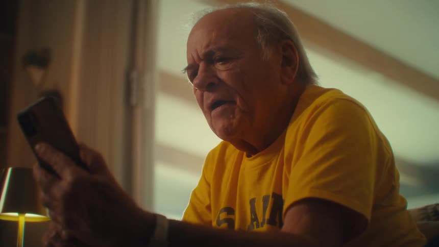 An elderly man wearing a yellow tshirt sitting in his living room reading a magazine suddenly picks up his mobile phone to answer a video call becoming overly excited as he waves to the caller while talking