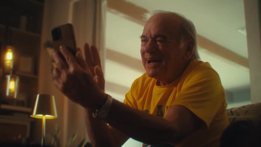 An elderly man sitting on a couch in his living room excited and smiling as he waves and talks during a video call on his mobile phone