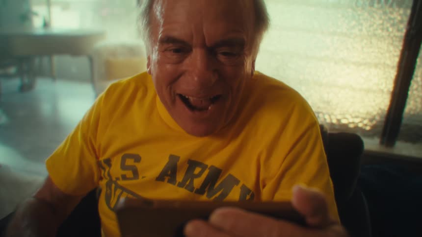 An elderly man beams with excitement while holding a phone wearing a bright yellow us army shirt
