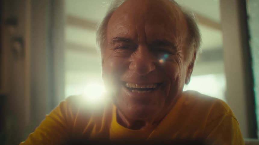 An elderly man sitting on a couch indoors laughing while watching content on his phone