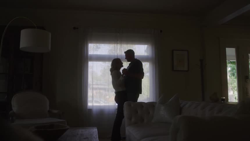 A couple hugging each other while standing beside a window in their living room at midday