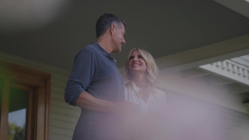 A couple stands on their front porch at midday smiling at each other as the man holds a cup of coffee