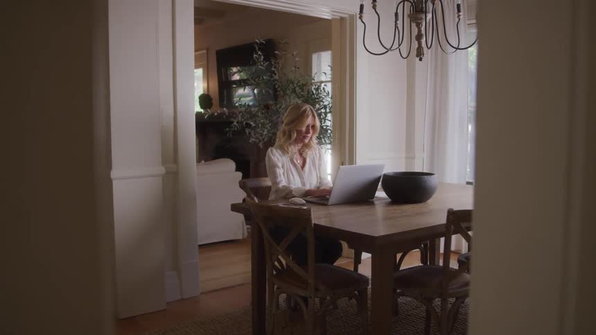 A woman works peacefully on a laptop at a wooden dining table in a sunlit home