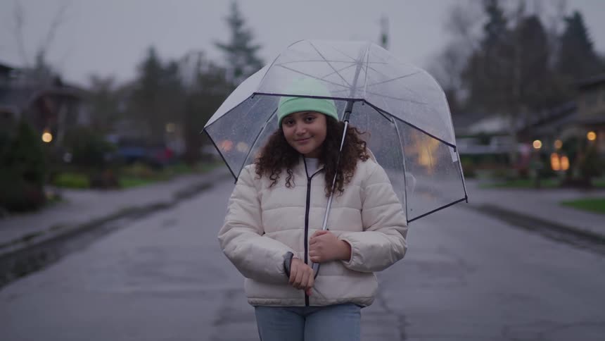 A teenager with curly brown hair and a mint green beanie stands on a wet street while holding a clear umbrella and looking at the camera